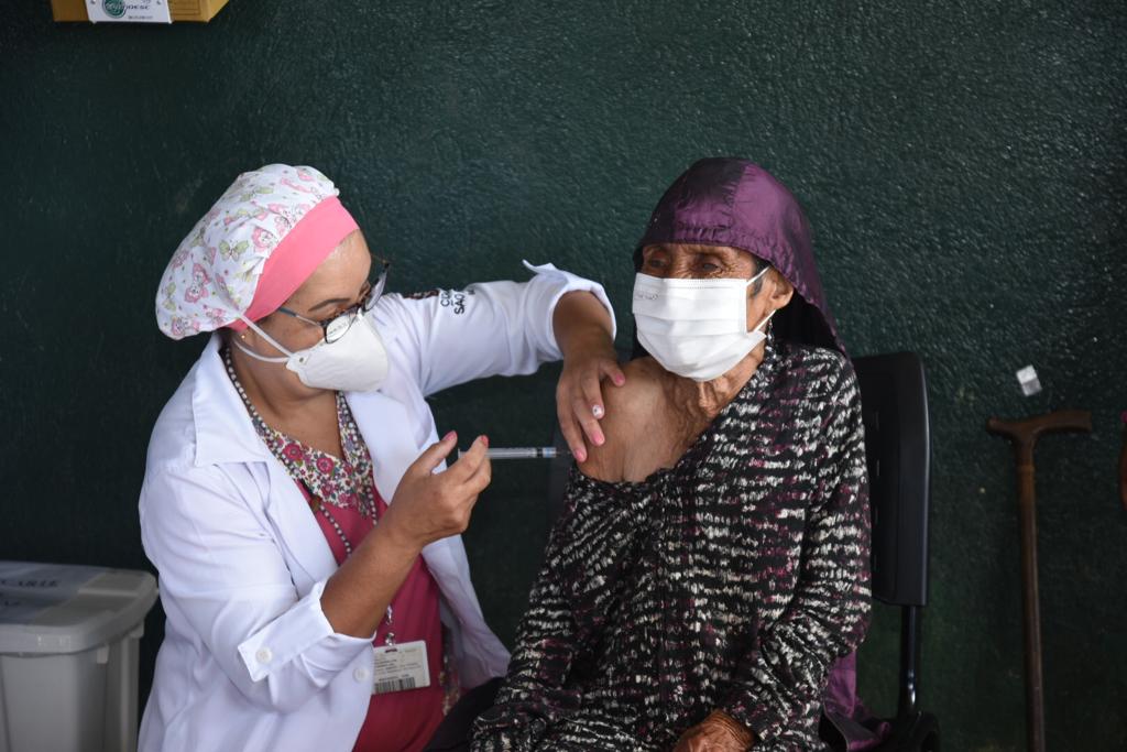 Sentada numa cadeira, a índia Maria Rita da Silva, 107 anos, com chapéu bordô e vestido estampado miúdo em preto e branco, com o ombro direito à mostra recebe a vacina de uma agente de saúde, que usa jaleco branco, touca rosa, máscara e óculos